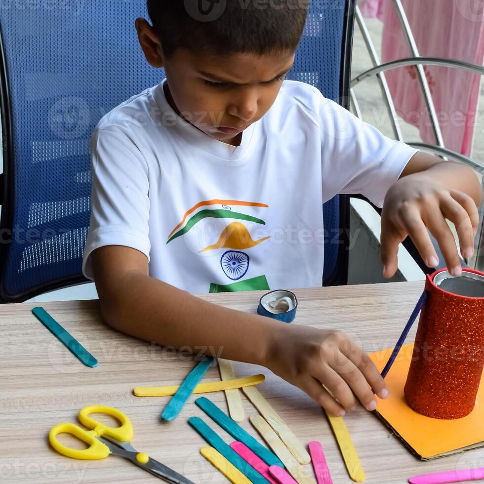 Smart Indian little boy performing best out of waste activity with different colourful objects during the summer vacations, Cute Indian Kid doing best our of waste activity at home on wooden table photo