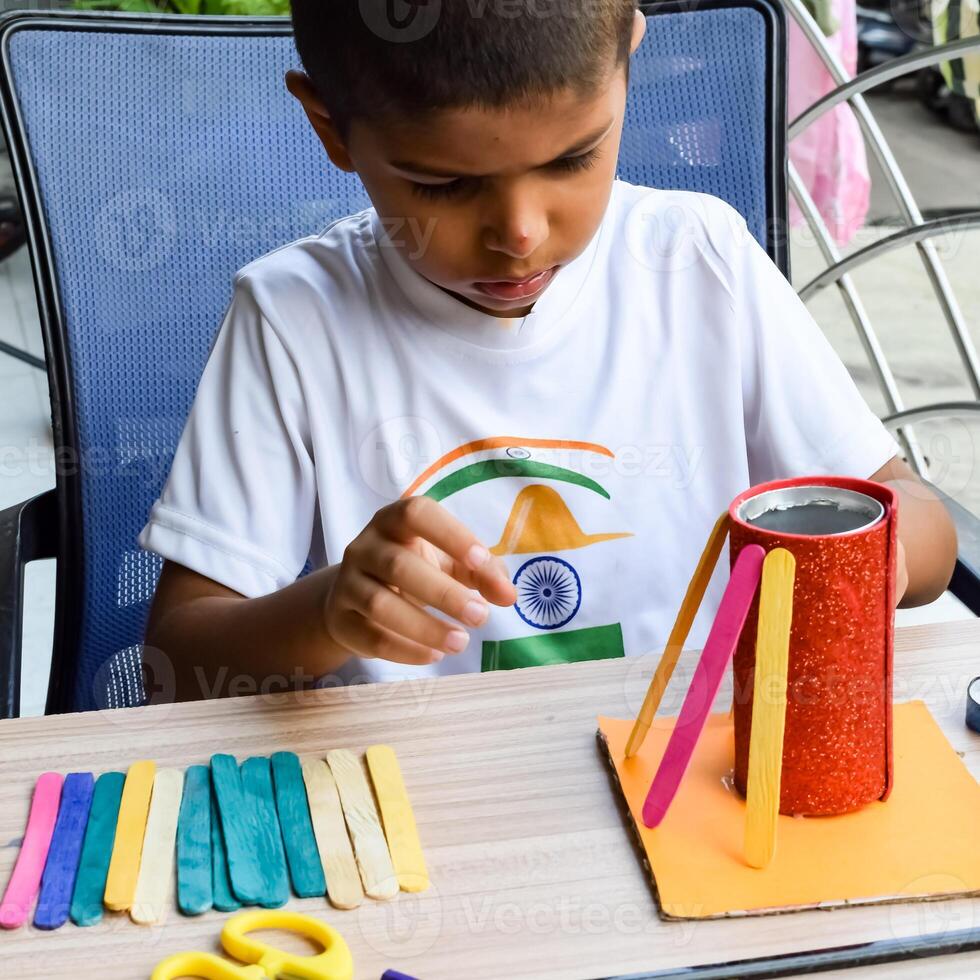 Smart Indian little boy performing best out of waste activity with different colourful objects during the summer vacations, Cute Indian Kid doing best our of waste activity at home on wooden table photo
