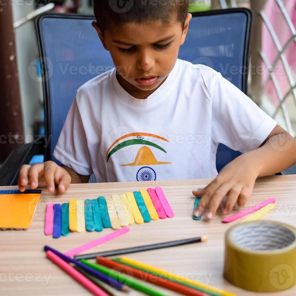 Smart Indian little boy performing best out of waste activity with different colourful objects during the summer vacations, Cute Indian Kid doing best our of waste activity at home on wooden table photo