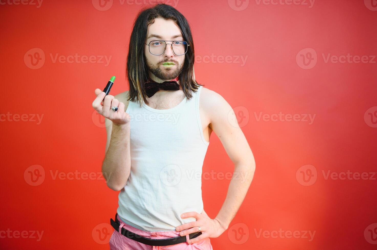 Young man applying lip gloss on his lips and looking in camera like mirror. Gay man doing make up against red background photo