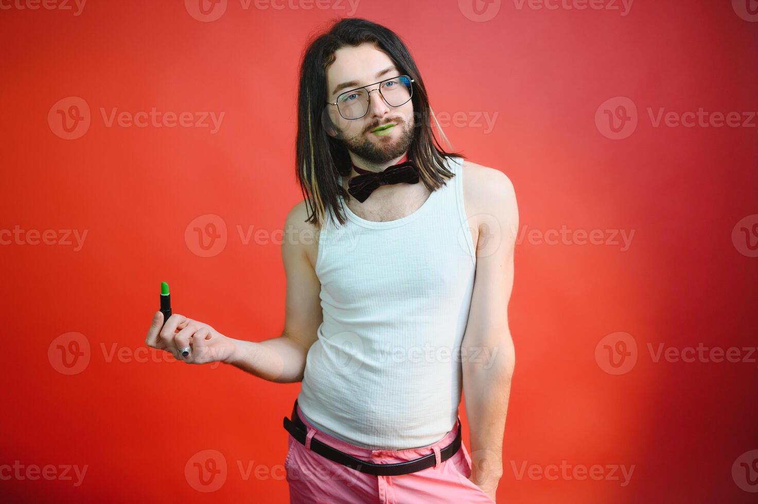 Young man applying lip gloss on his lips and looking in camera like mirror. Gay man doing make up against red background photo