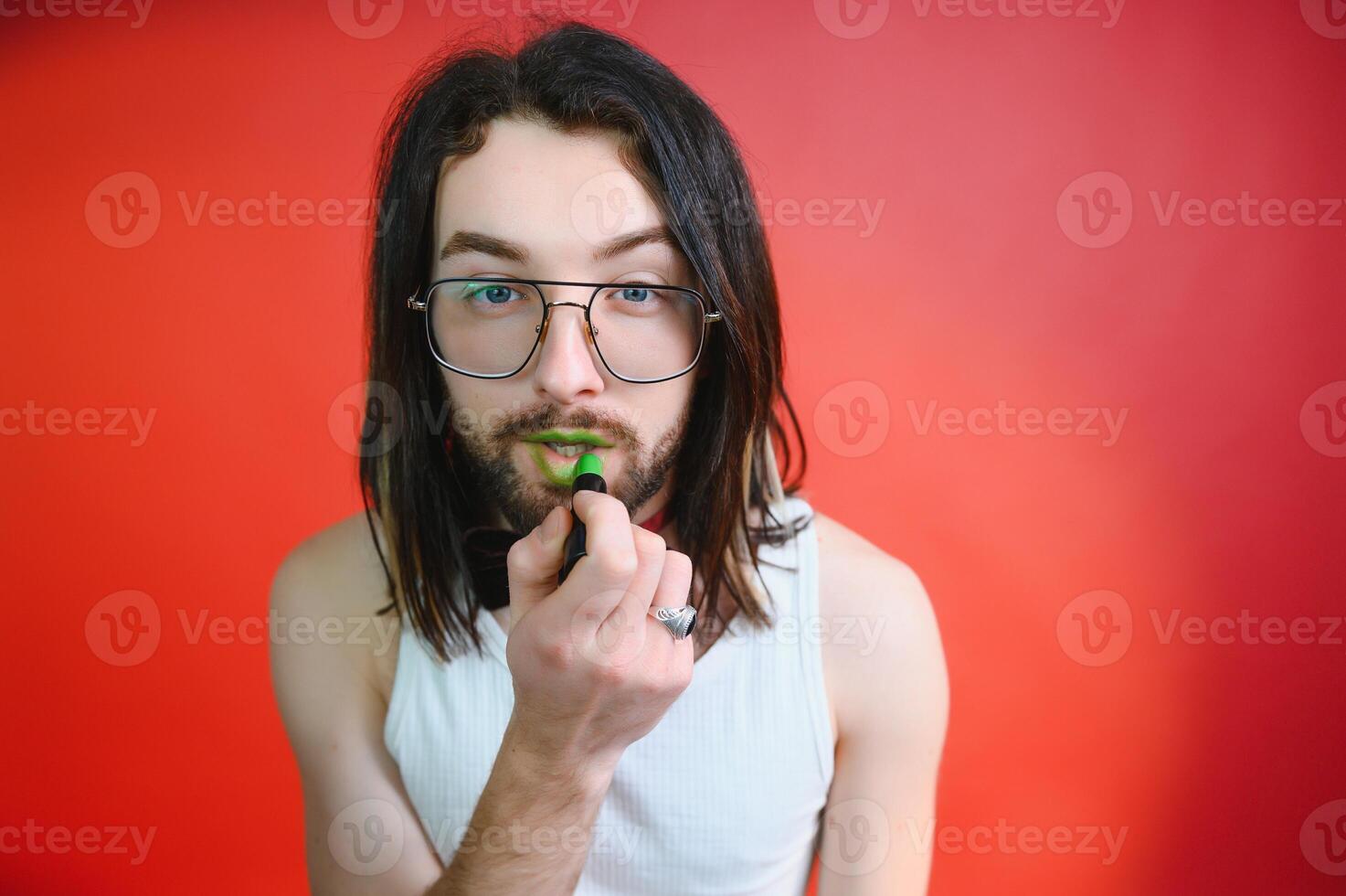 Young man applying lip gloss on his lips and looking in camera like mirror. Gay man doing make up against red background photo