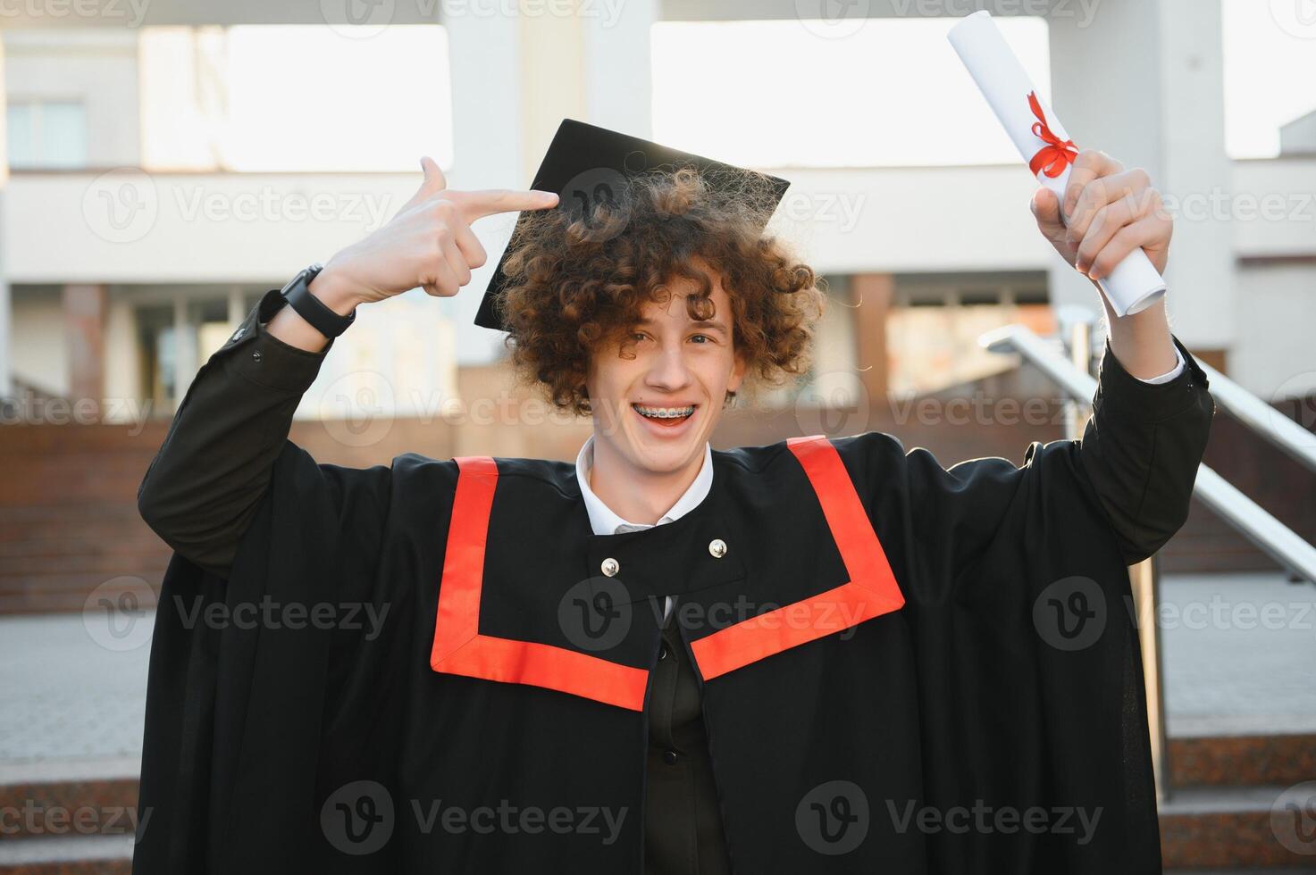 Graduation from university. Young smiling boy university graduate in traditional bonet and mantle standing and holding diploma in hand over university building background photo