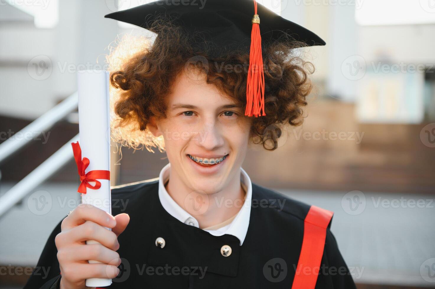 Graduation from university. Young smiling boy university graduate in traditional bonet and mantle standing and holding diploma in hand over university building background photo