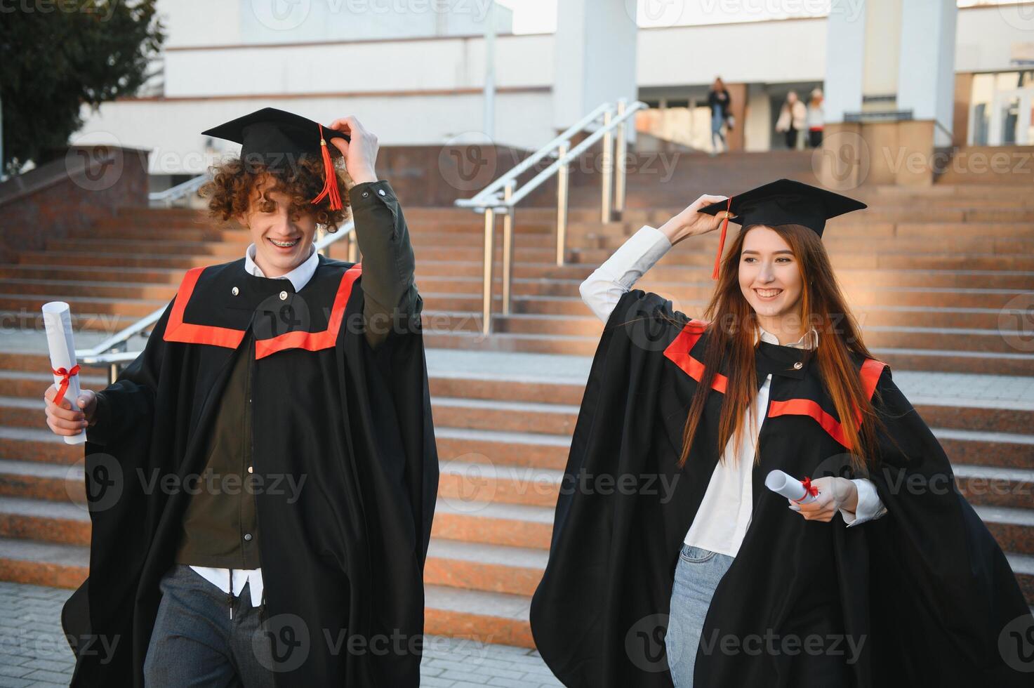Portrait of happy graduates. Two friends in graduation caps and gowns standing outside university building with other students in background, holding diploma scrolls, and smiling photo