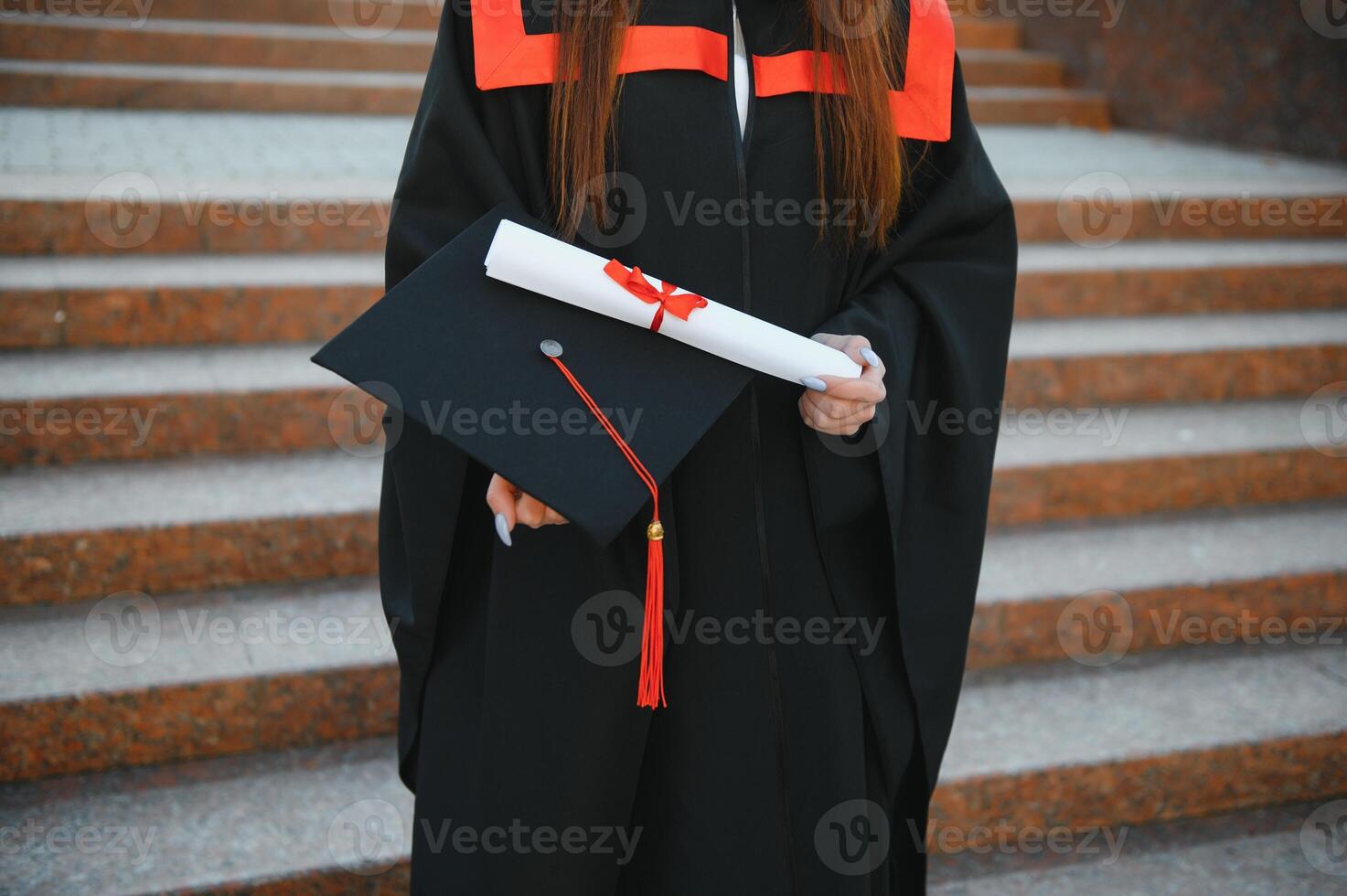 Happy cute brunette caucasian grad girl is smiling. She is in a black mortar board, with red tassel, in gown, with nice brown curly hair, diploma in hand. photo