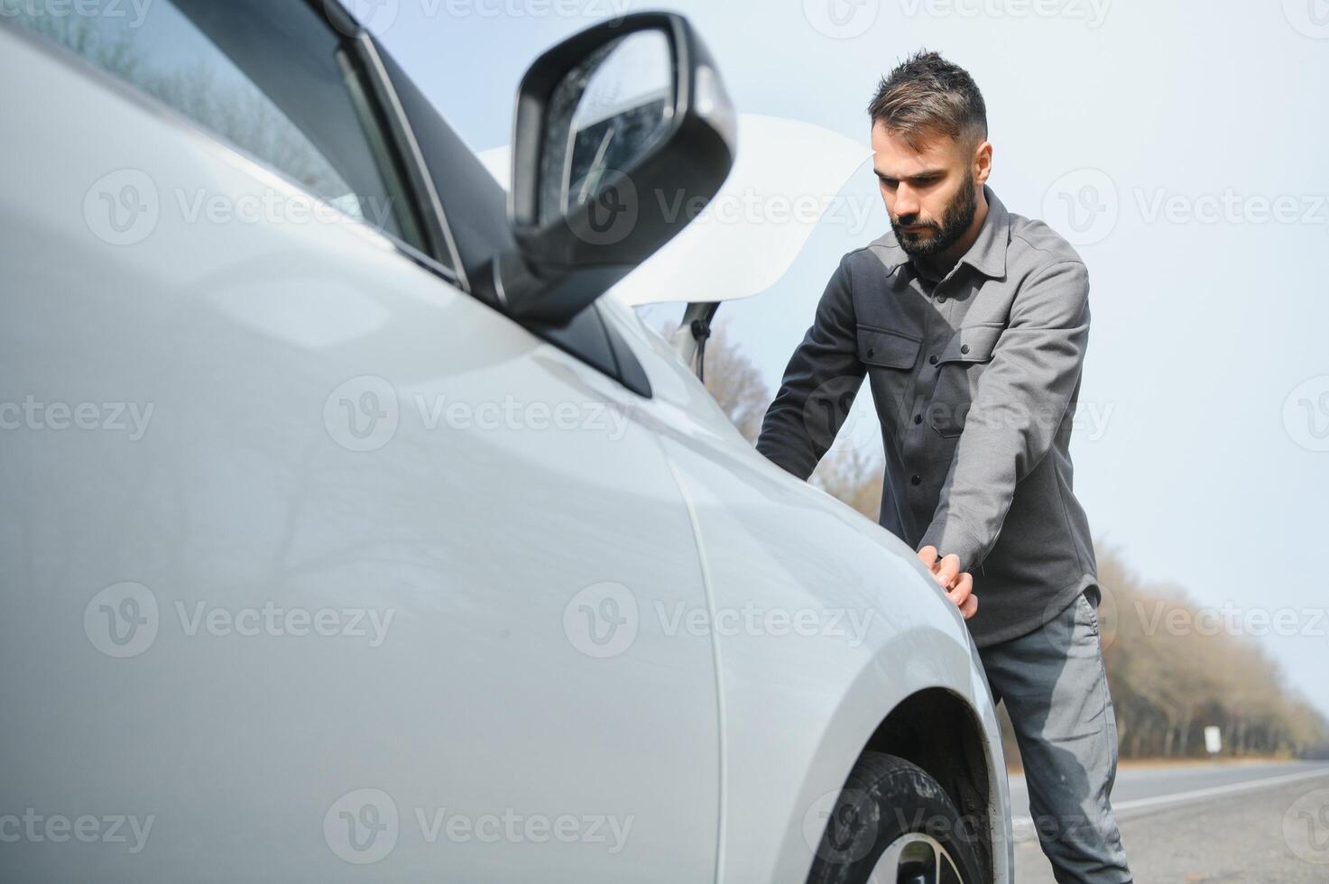 A young man near a broken car with an open hood on the roadside. photo