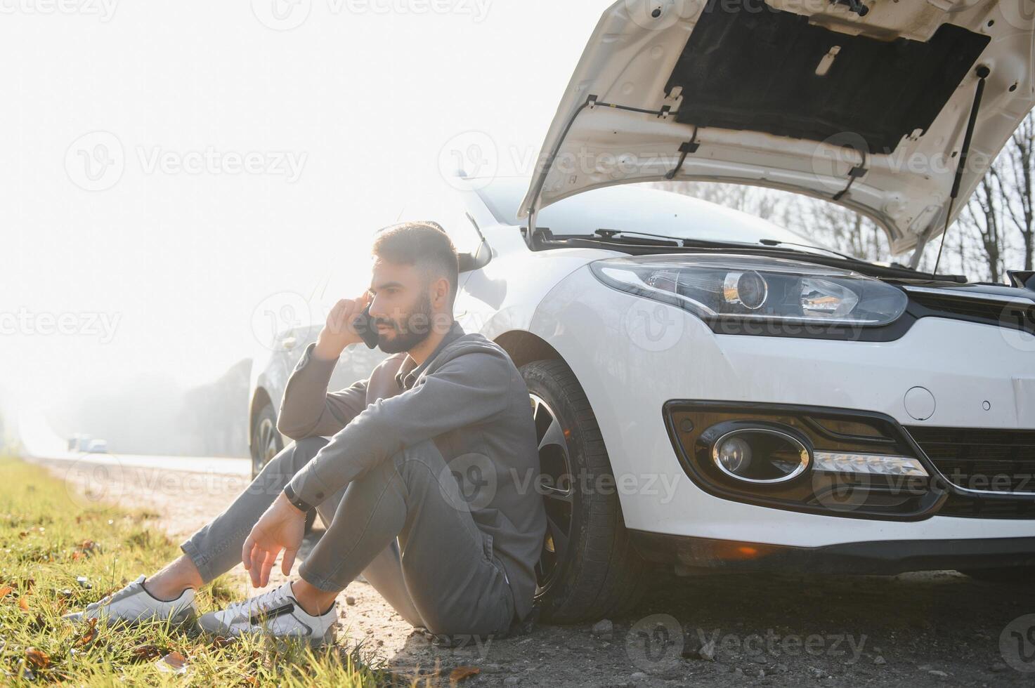 Picture of frustrated man sitting next to broken car with open hood photo