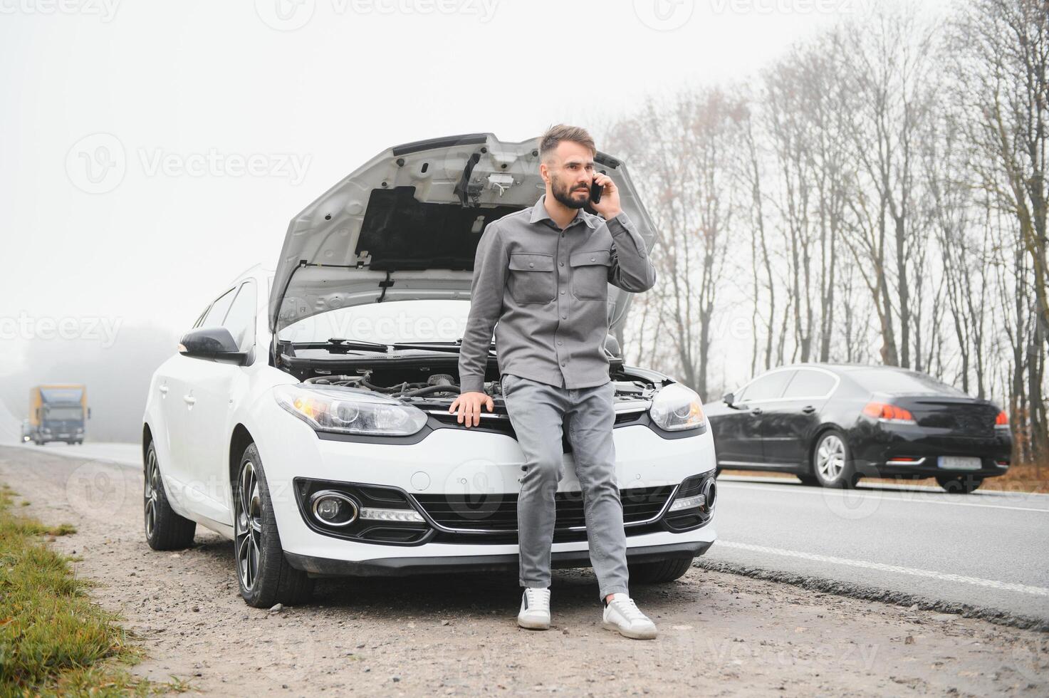 A young man near a broken car with an open hood on the roadside. photo