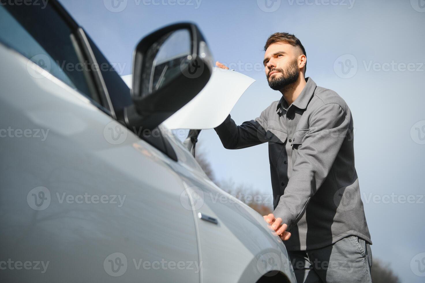 Young upset casual man trying to fix his broken car outdoors. Man waiting for towing service for help car accident on the road. Roadside assistance concept. photo