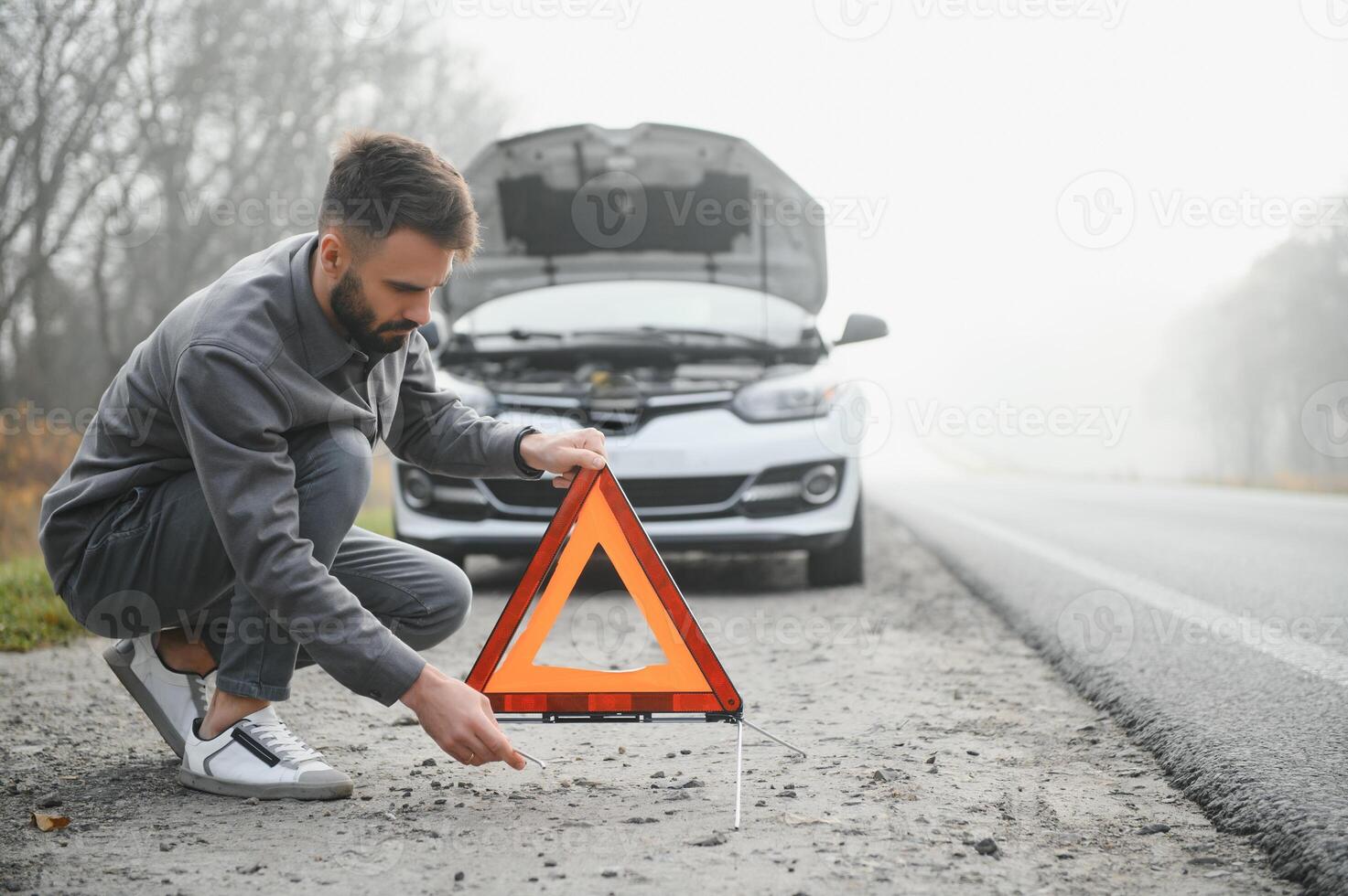 A young man near a broken car with an open hood on the roadside. photo