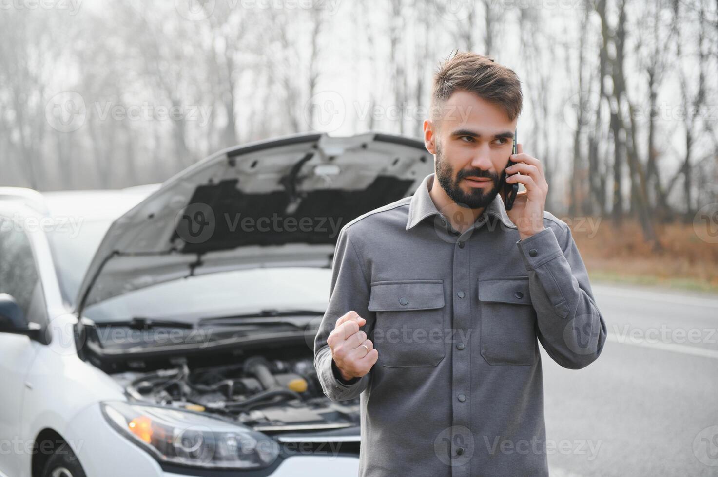A young man near a broken car with an open hood on the roadside. photo