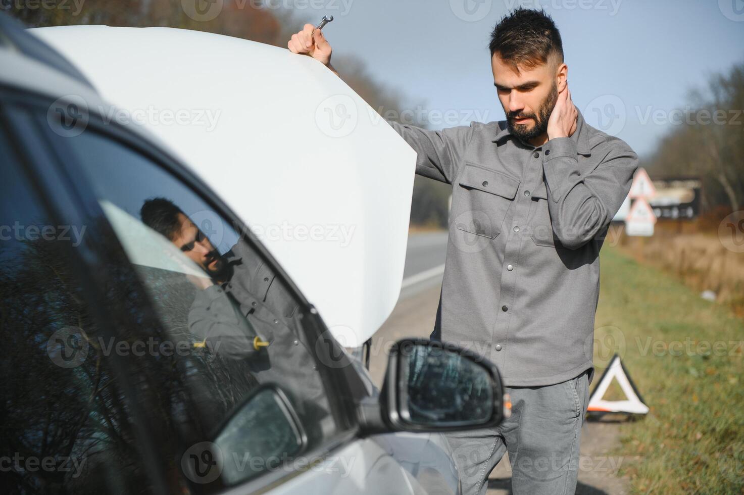 A young man near a broken car with an open hood on the roadside. photo