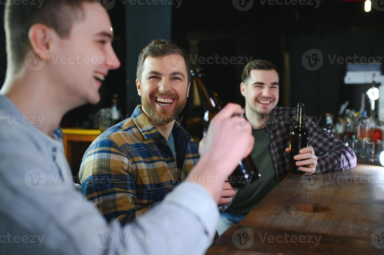 Three young men in casual clothes are smiling, holding bottles of beer while standing near bar counter in pub photo