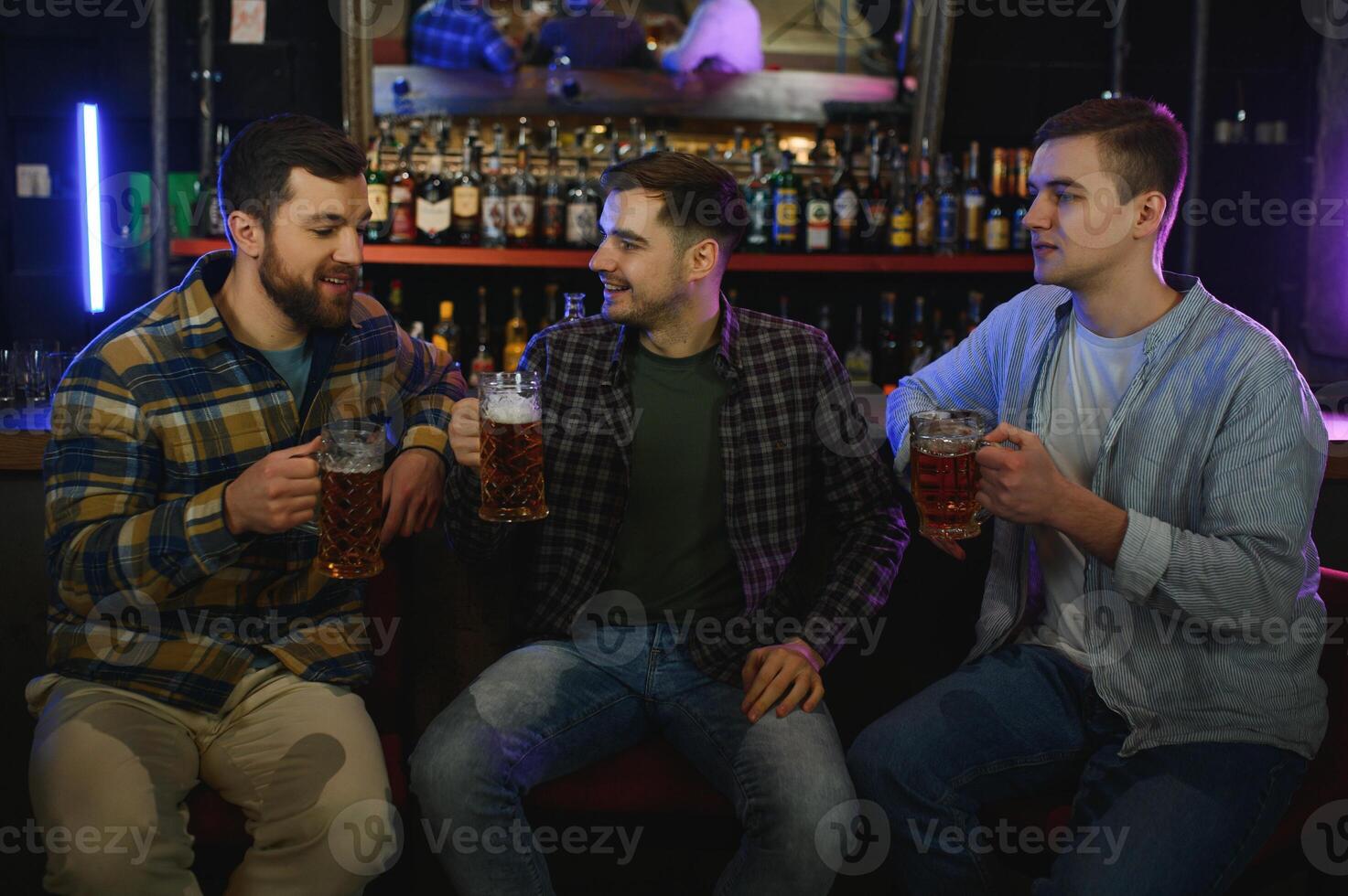Three young men in casual clothes are smiling, holding bottles of beer while standing near bar counter in pub photo