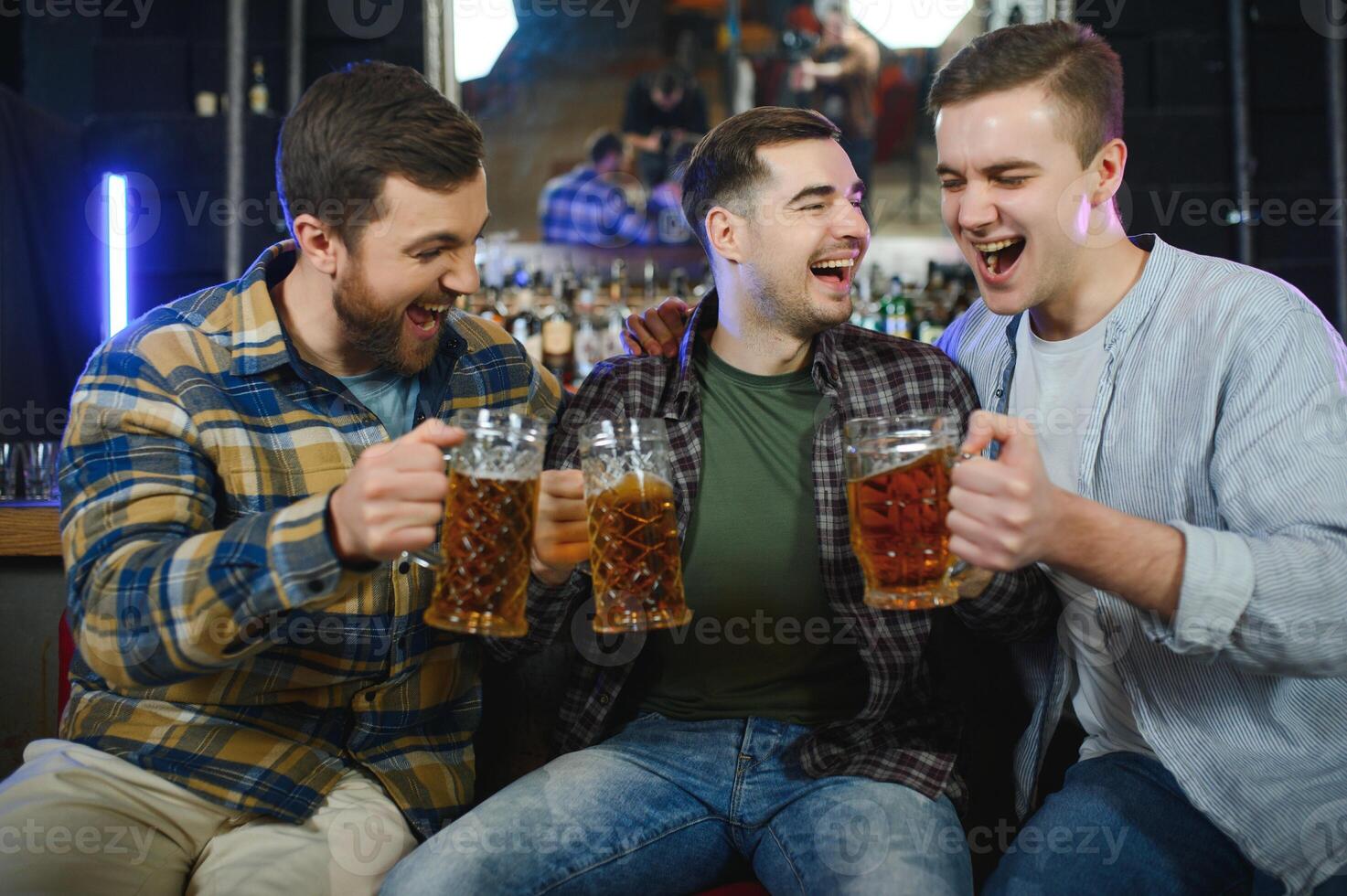 Three young men in casual clothes are smiling and clanging glasses of beer together while sitting at bar counter in pub photo