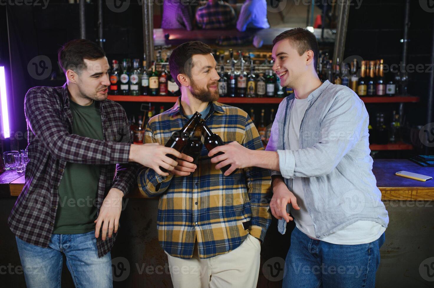 Three young men in casual clothes are smiling and clanging glasses of beer together while sitting at bar counter in pub photo