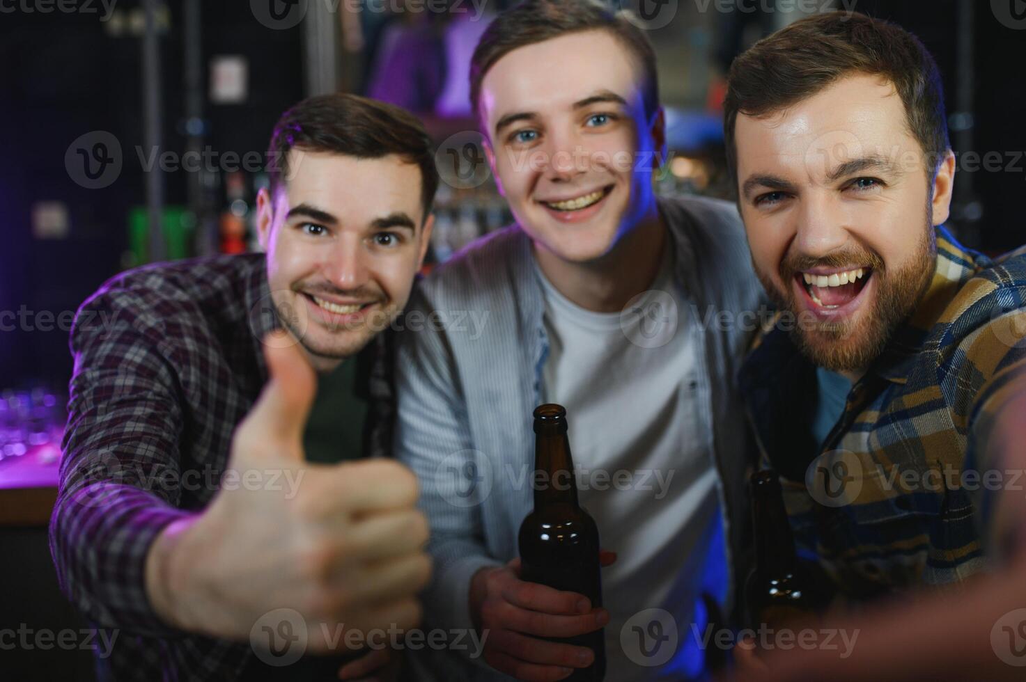 Three young men in casual clothes are smiling, holding bottles of beer while standing near bar counter in pub photo