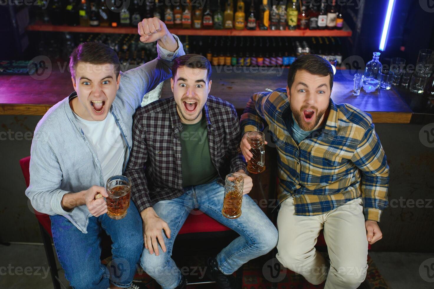 Soccer fans cheering for favourite team while enjoying drinks. Group of supporters celebrating victory while watching football match on tv in a club. Happy friends shouting while cheering for a goal. photo