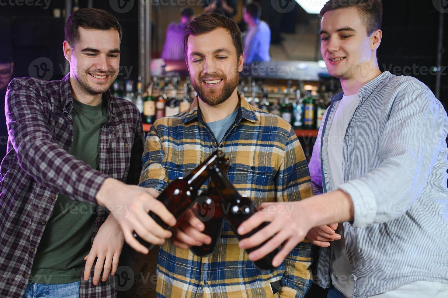 Three young men in casual clothes are smiling, holding bottles of beer while standing near bar counter in pub photo