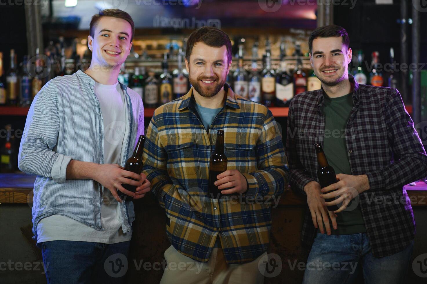 Three young men in casual clothes are smiling, holding bottles of beer while standing near bar counter in pub photo