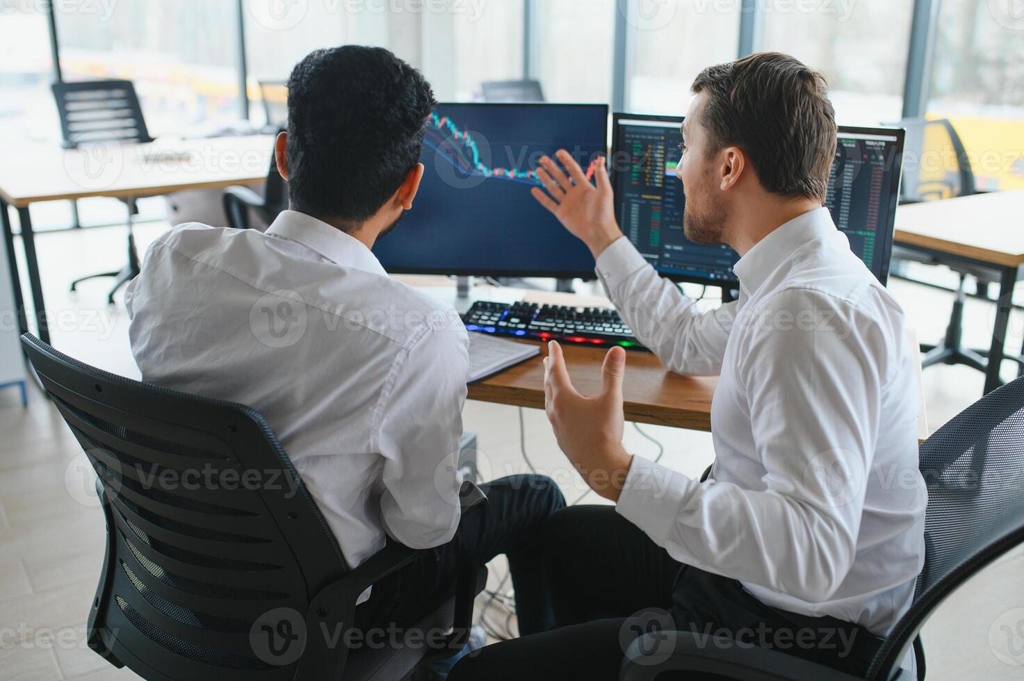 Two men traders sitting at desk at office together monitoring stocks data candle charts on screen analyzing price flow smiling cheerful having profit teamwork concept photo