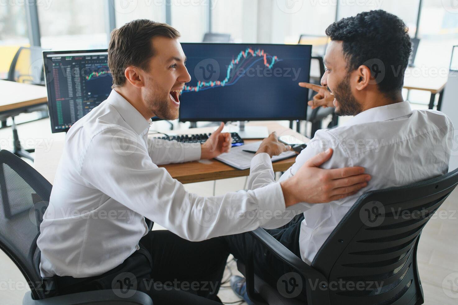 Two men traders sitting at desk at office together monitoring stocks data candle charts on screen analyzing price flow smiling cheerful having profit teamwork concept photo