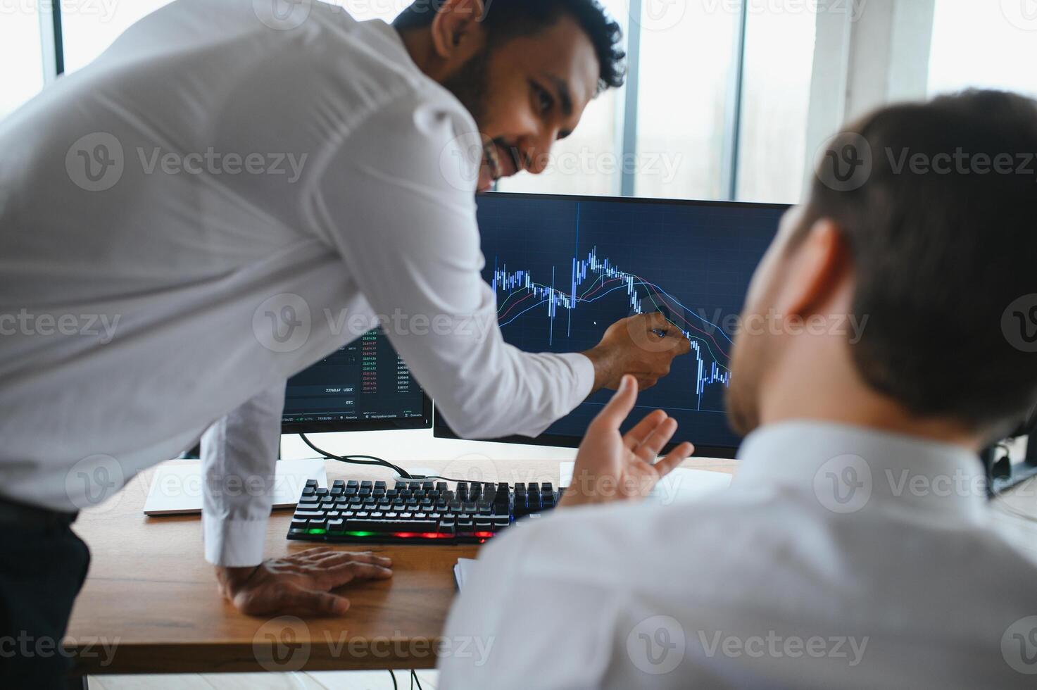 Two successful trader sitting in office and looking at screen, checking cryptocurrency information data on finance market graph, pointing on monitor photo