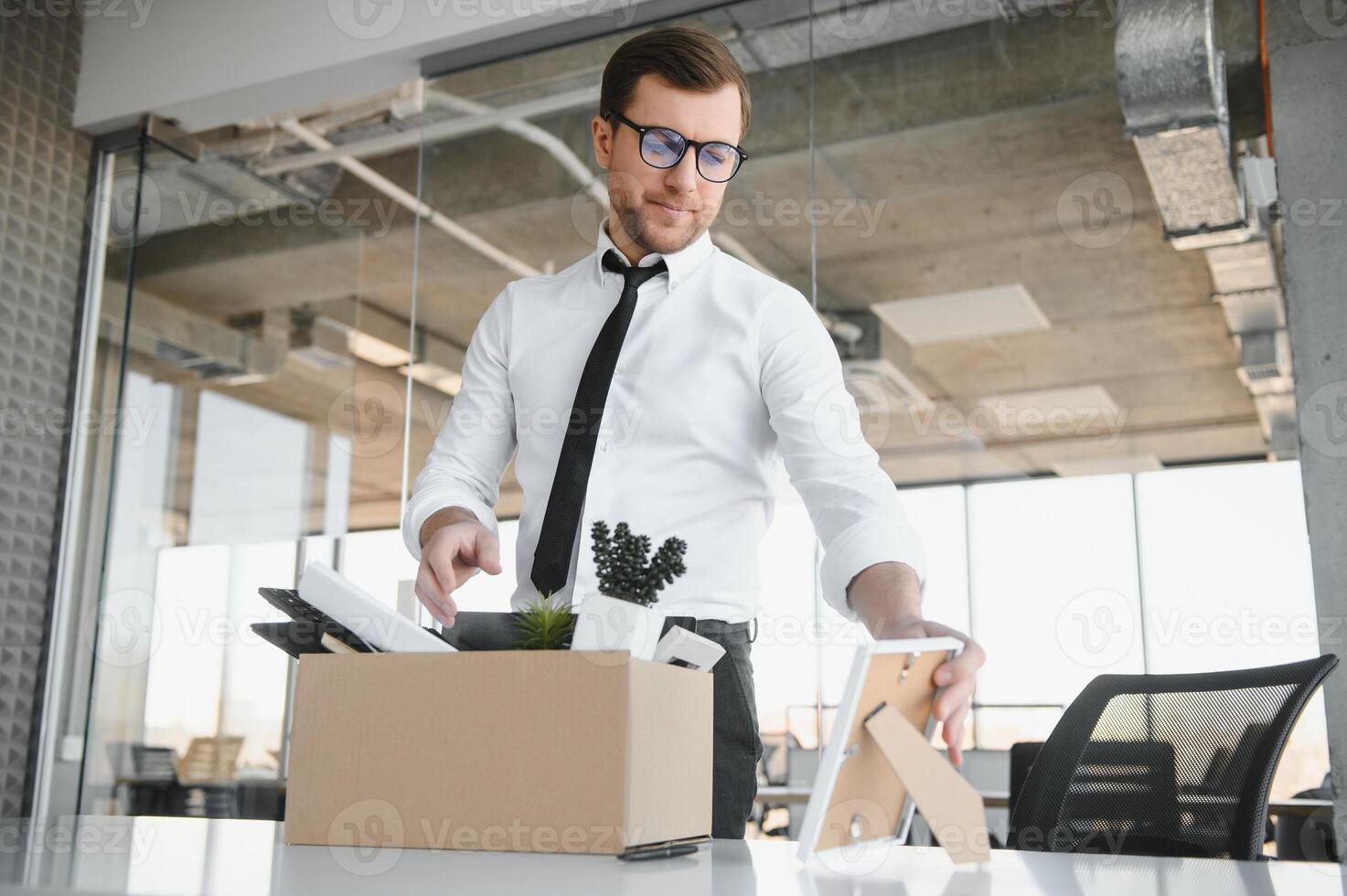 Young handsome businessman in light modern office with carton box. Last day at work. Upset office worker is fired. photo