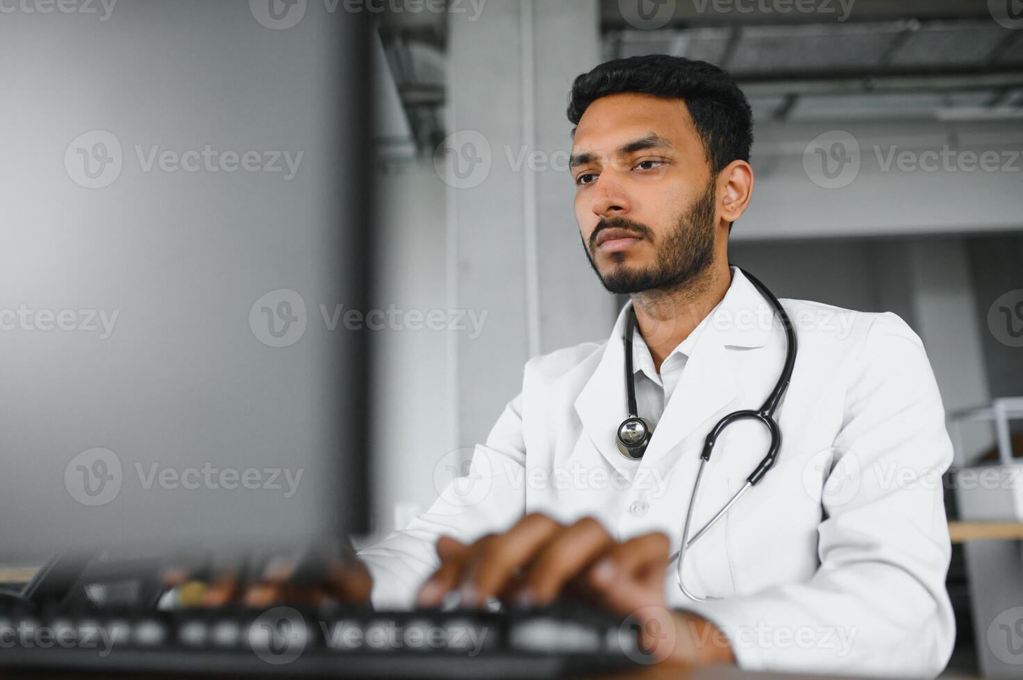 People, occupation and medicine concept. Smiling male Indian Arab doctor in white coat, sitting in medical office at desk with laptop photo