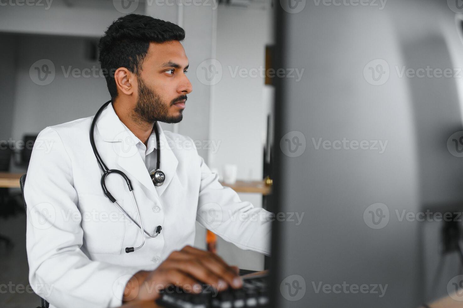 Indian male doctor physician wearing glasses, white medical gown and stethoscope sitting at the desk with the laptop in modern clinic and involved online onference, consulting remotely. photo