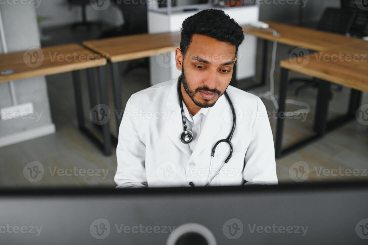 People, occupation and medicine concept. Smiling male Indian Arab doctor in white coat, sitting in medical office at desk with laptop photo