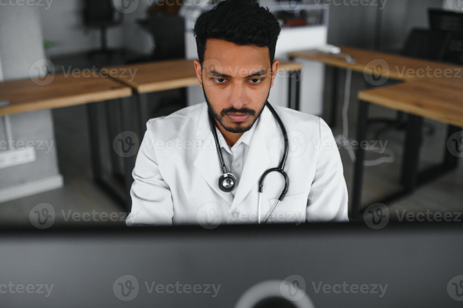 Indian male doctor physician wearing glasses, white medical gown and stethoscope sitting at the desk with the laptop in modern clinic and involved online onference, consulting remotely. photo