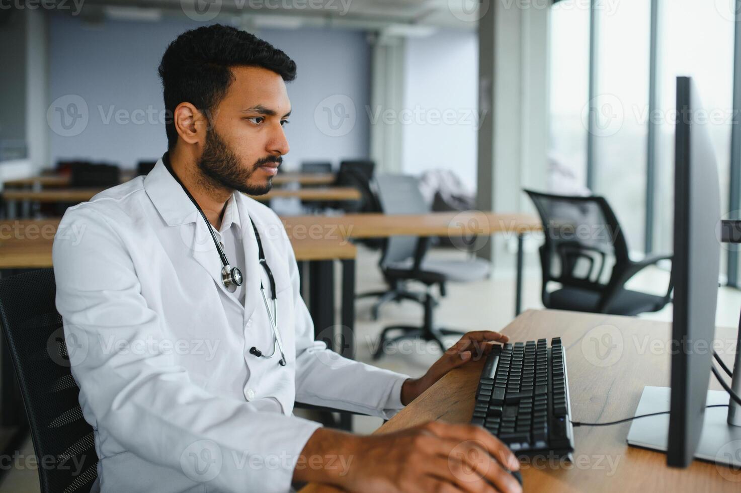 People, occupation and medicine concept. Smiling male Indian Arab doctor in white coat, sitting in medical office at desk with laptop photo