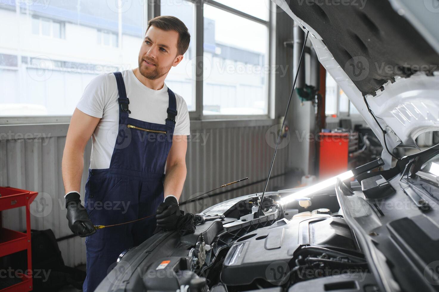 Handsome young male auto mechanic in special uniform clothes holding a flashlight, looking for breakdown and repairing under the hood in the car engine in a car workshop photo