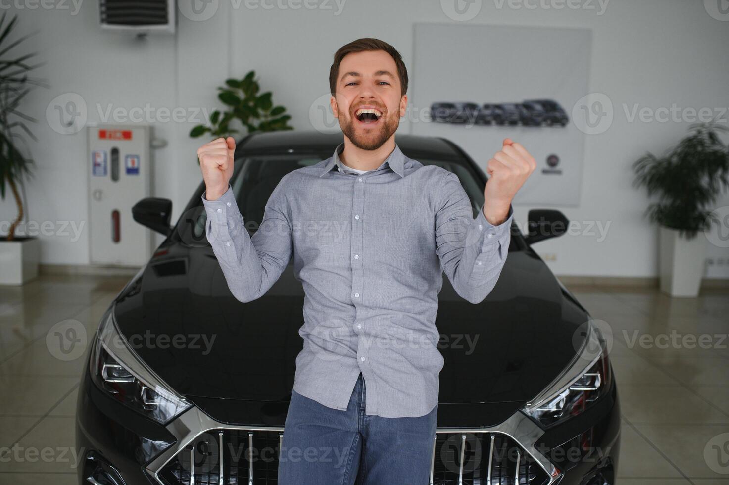 Emotional handsome guy raising hand up and smiling at camera. Rich man buying auto at luxury dealership salon photo