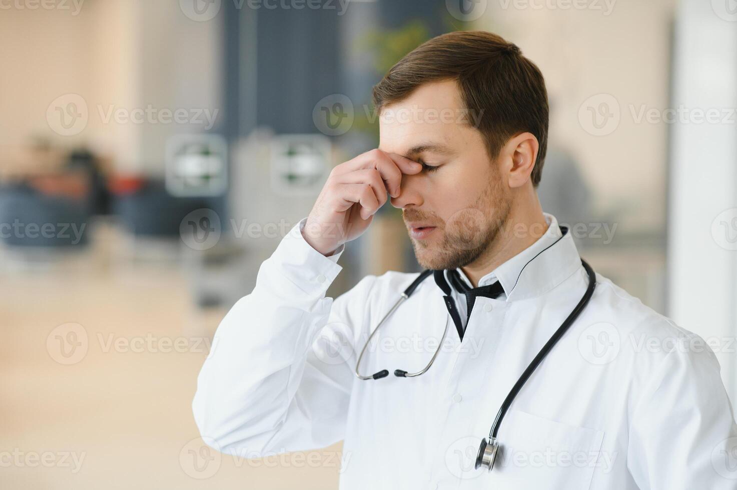 Handsome young doctor in white coat is touching his temples while standing in hospital hall. photo