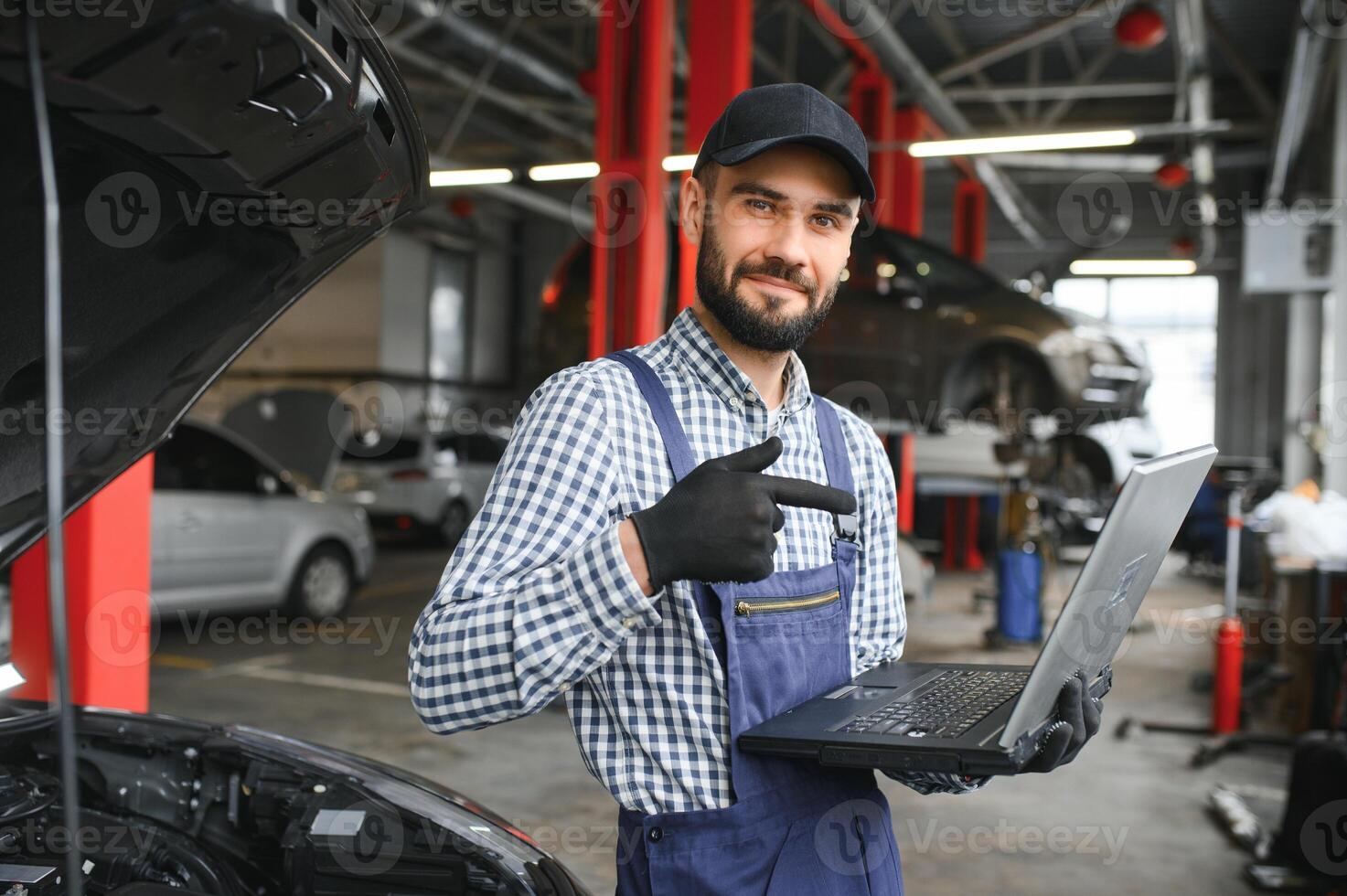 A bearded auto-mechanic in working uniform is standing next to a broken car and using the special laptop for car maintenance. An auto-mechanic in workshop repairs car. photo