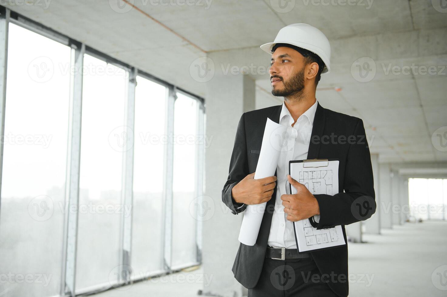Indian construction site manager standing wearing helmet, thinking at construction site. Portrait of mixed race manual worker or architect. photo