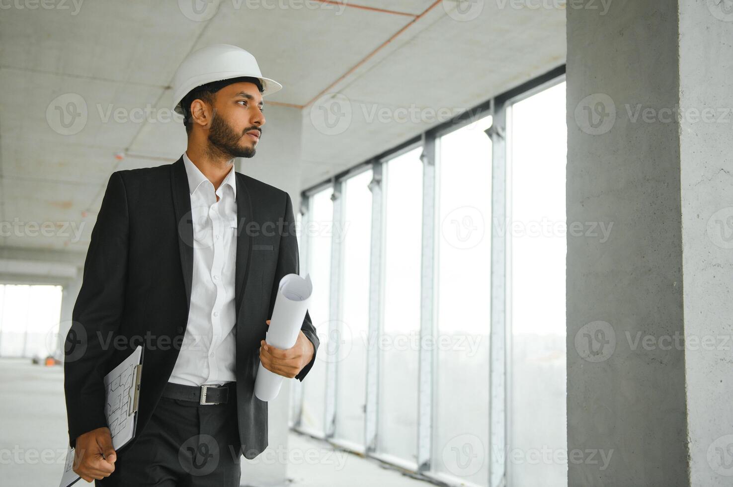 Indian construction site manager standing wearing helmet, thinking at construction site. Portrait of mixed race manual worker or architect. photo