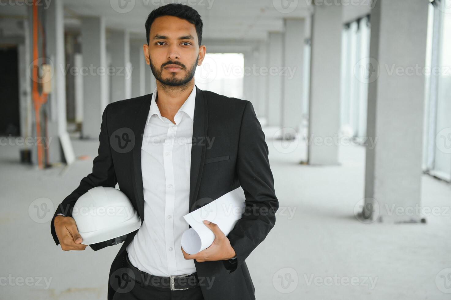 Indian construction site manager standing wearing helmet, thinking at construction site. Portrait of mixed race manual worker or architect. photo