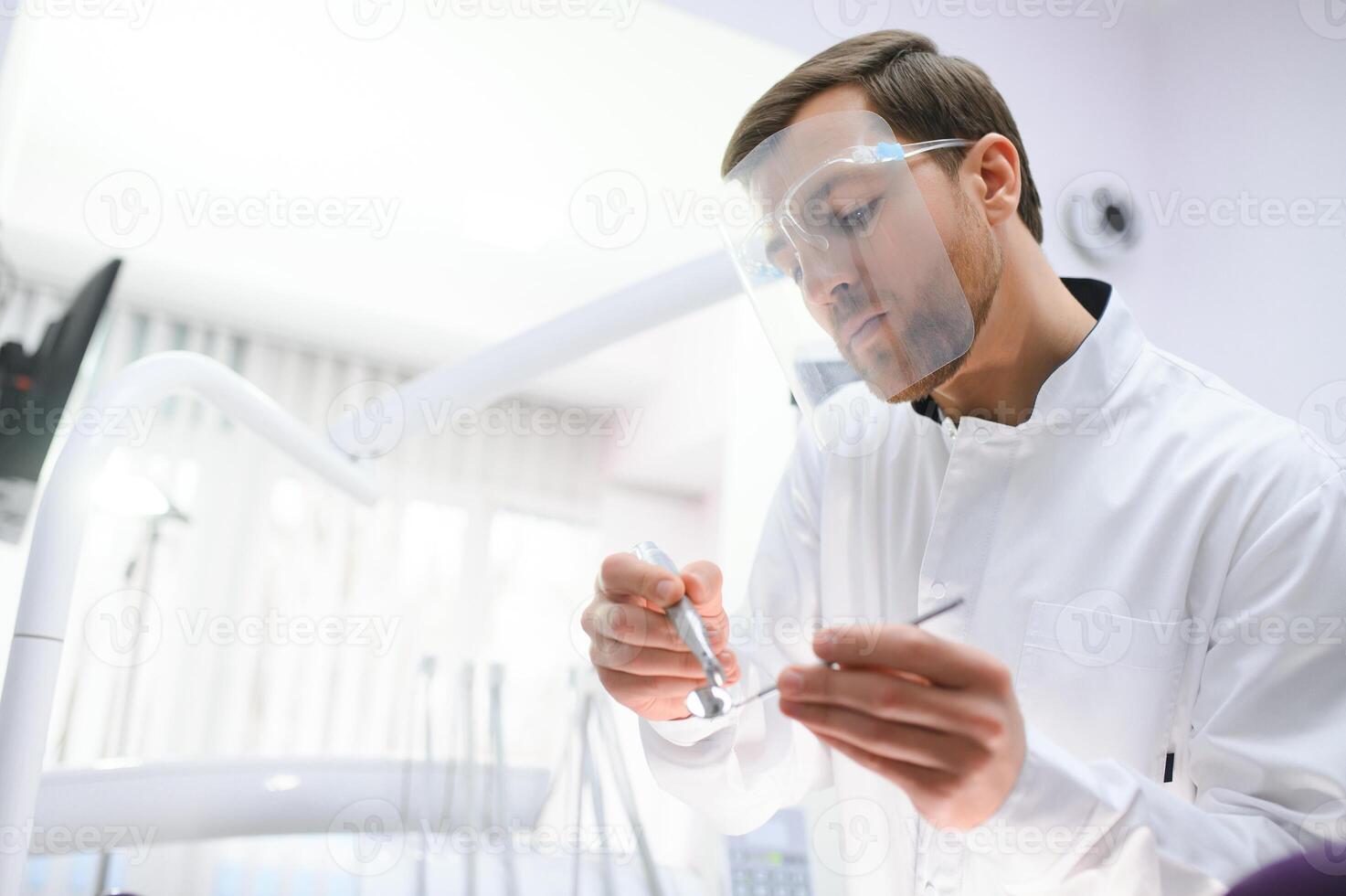 Bottom view young confident happy professional stomatologist european doctor man 20s in white gown hold showing tooth drilling machine at dentist office indoor cabinet. Healthcare aesthetic treatment. photo