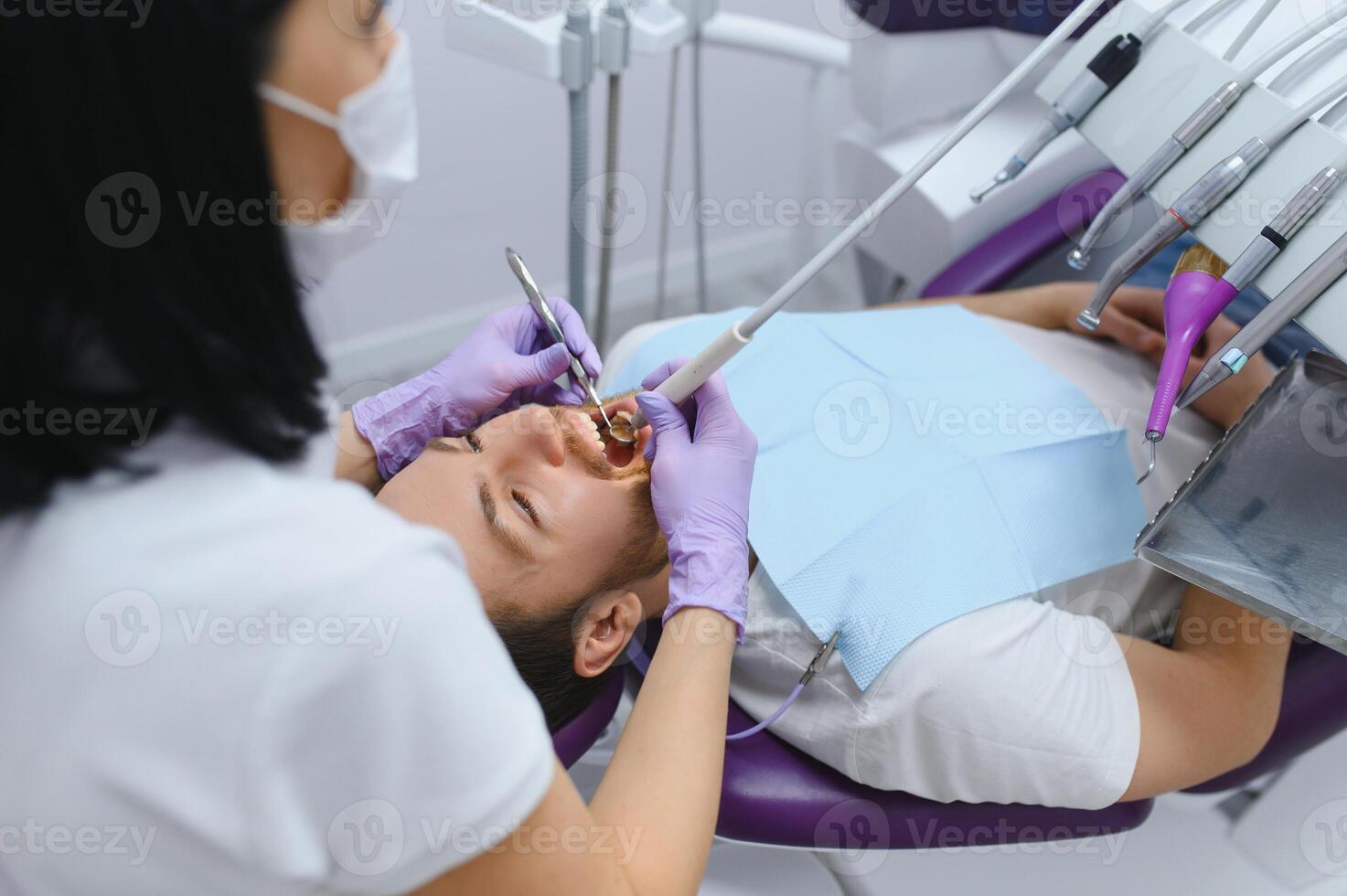 young bearded man sitting in dental chair, visiting modern dental clinic, having regular checkup, woman dentist. photo