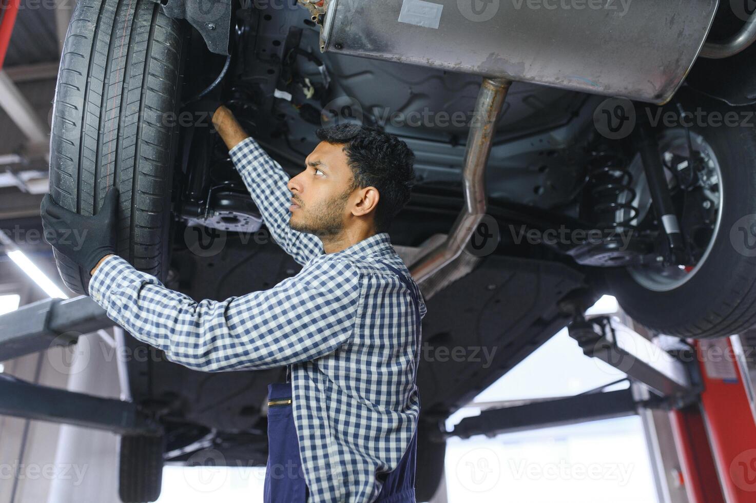 Indian car mechanic standing and working in service station. Car specialists examining the lifted car. Professional repairmen wearing mechanic uniform in blue color. photo
