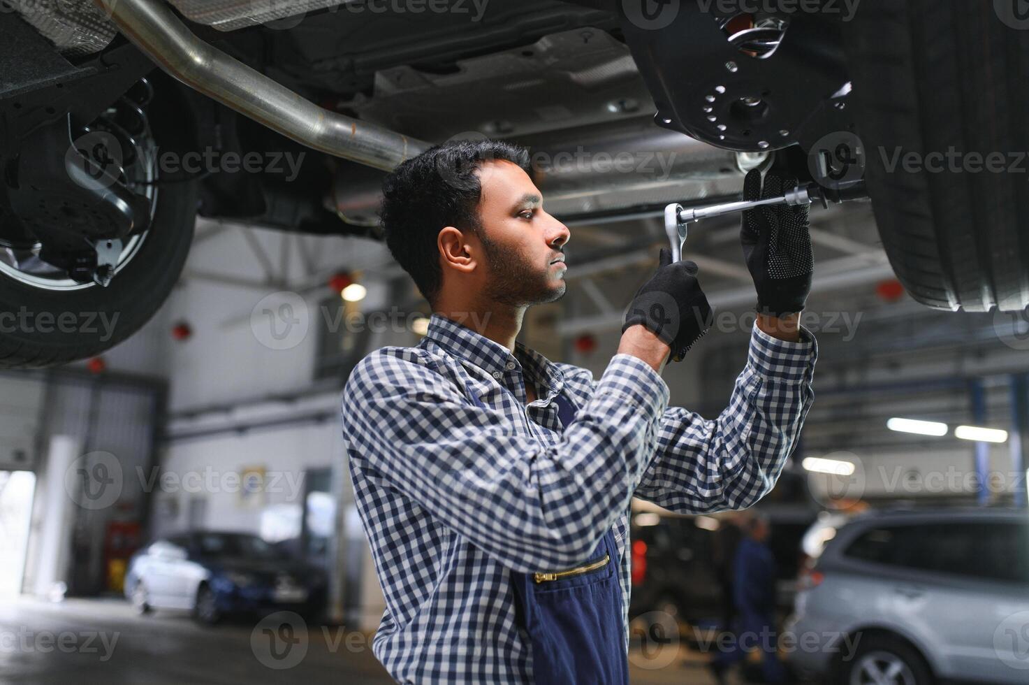 Indian car mechanic standing and working in service station. Car specialists examining the lifted car. Professional repairmen wearing mechanic uniform in blue color. photo