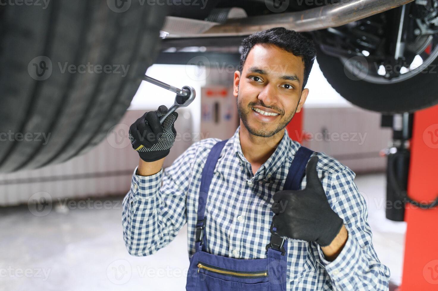 Indian car mechanic standing and working in service station. Car specialists examining the lifted car. Professional repairmen wearing mechanic uniform in blue color. photo