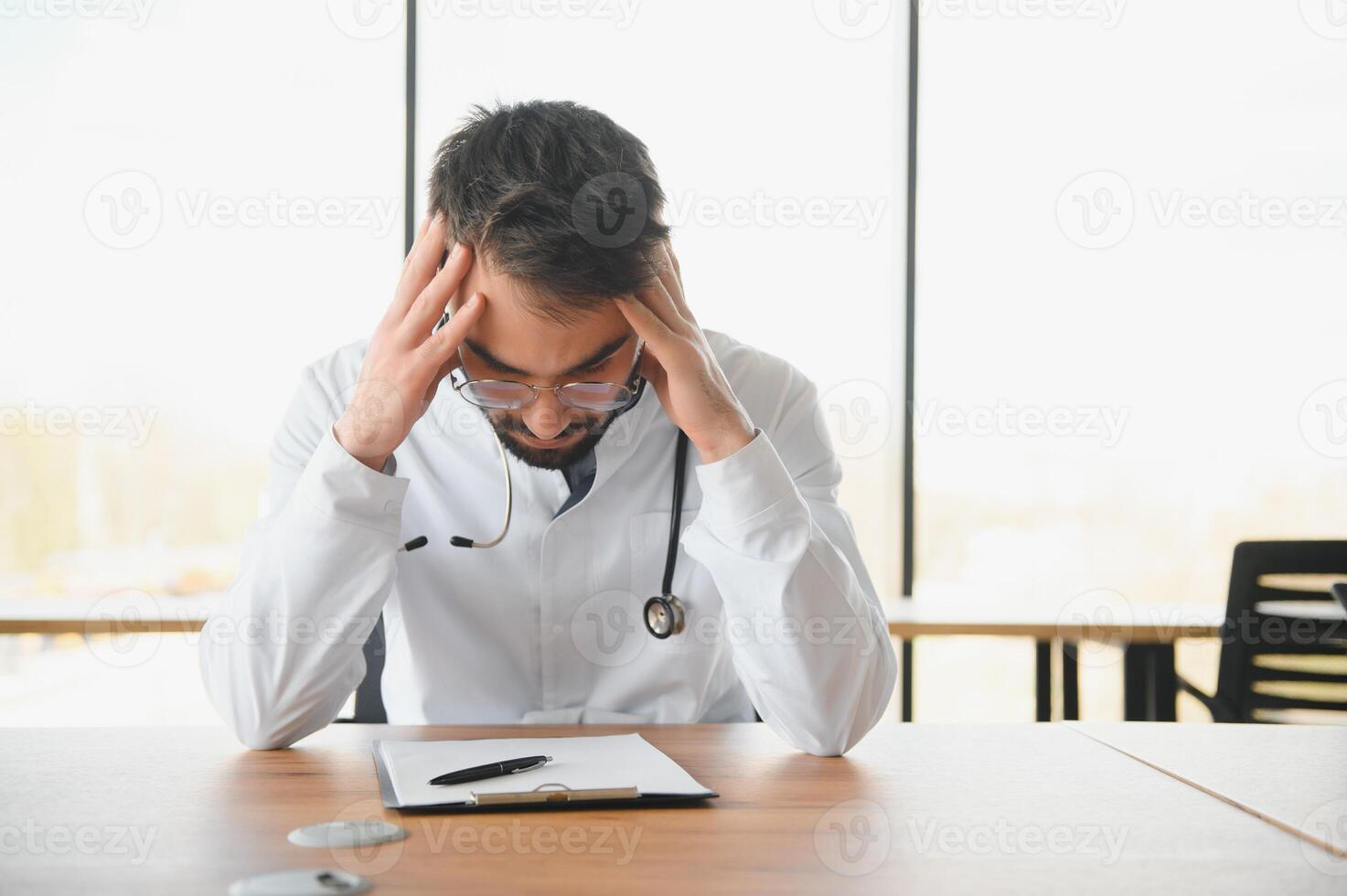 Handsome doctor man wearing medical uniform sitting on his workplace tired holding his head feeling fatigue and headache. Stress and frustration concept photo