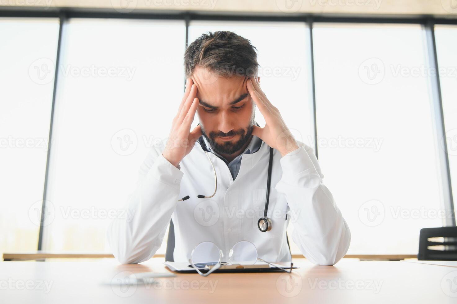 Close-up of upset tired medical worker closing head with hands. Depressed male doctor sitting in office. stethoscope on persons neck. Modern medicine and healthcare concept. photo