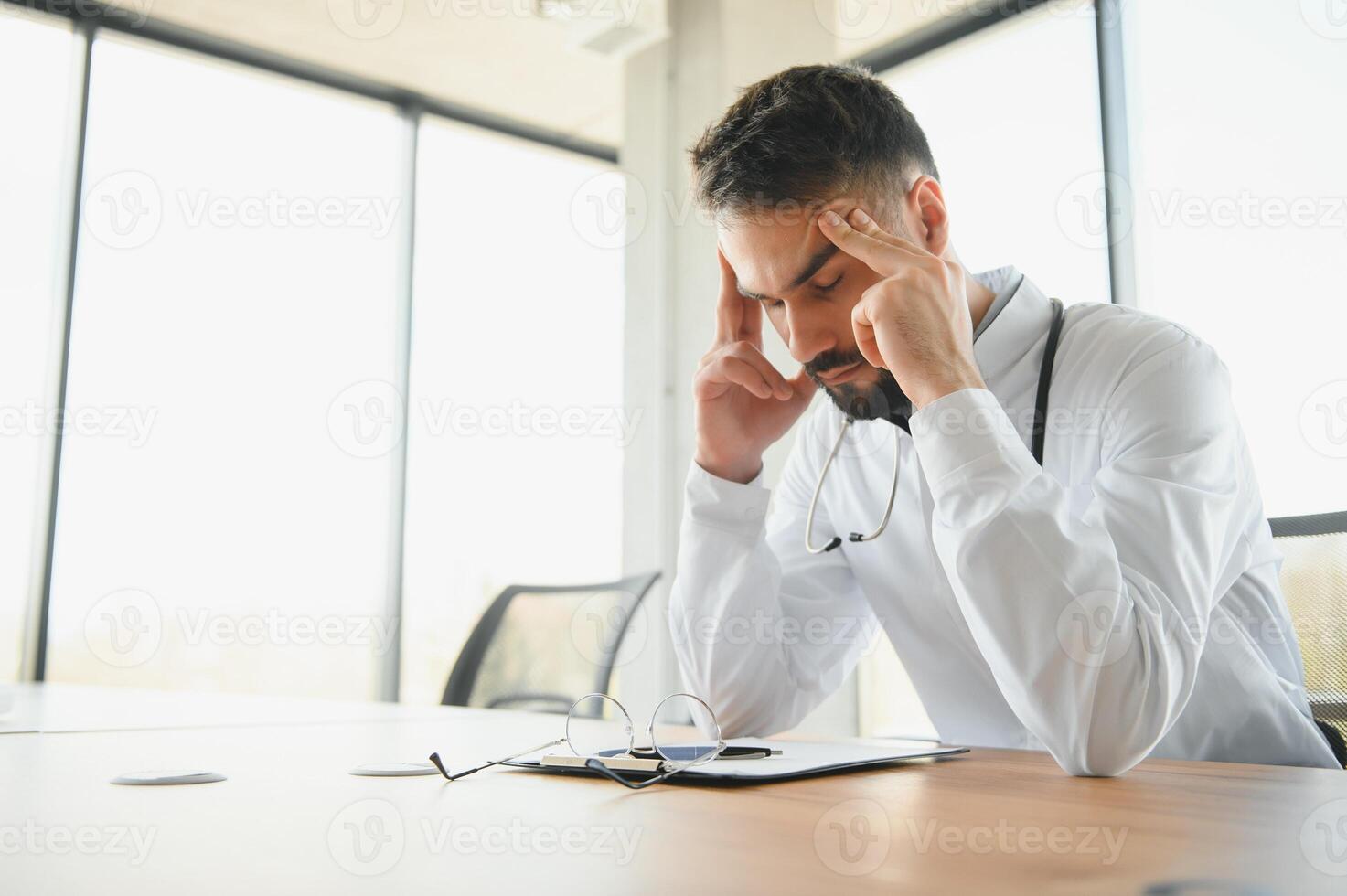 Close-up of upset tired medical worker closing head with hands. Depressed male doctor sitting in office. stethoscope on persons neck. Modern medicine and healthcare concept. photo