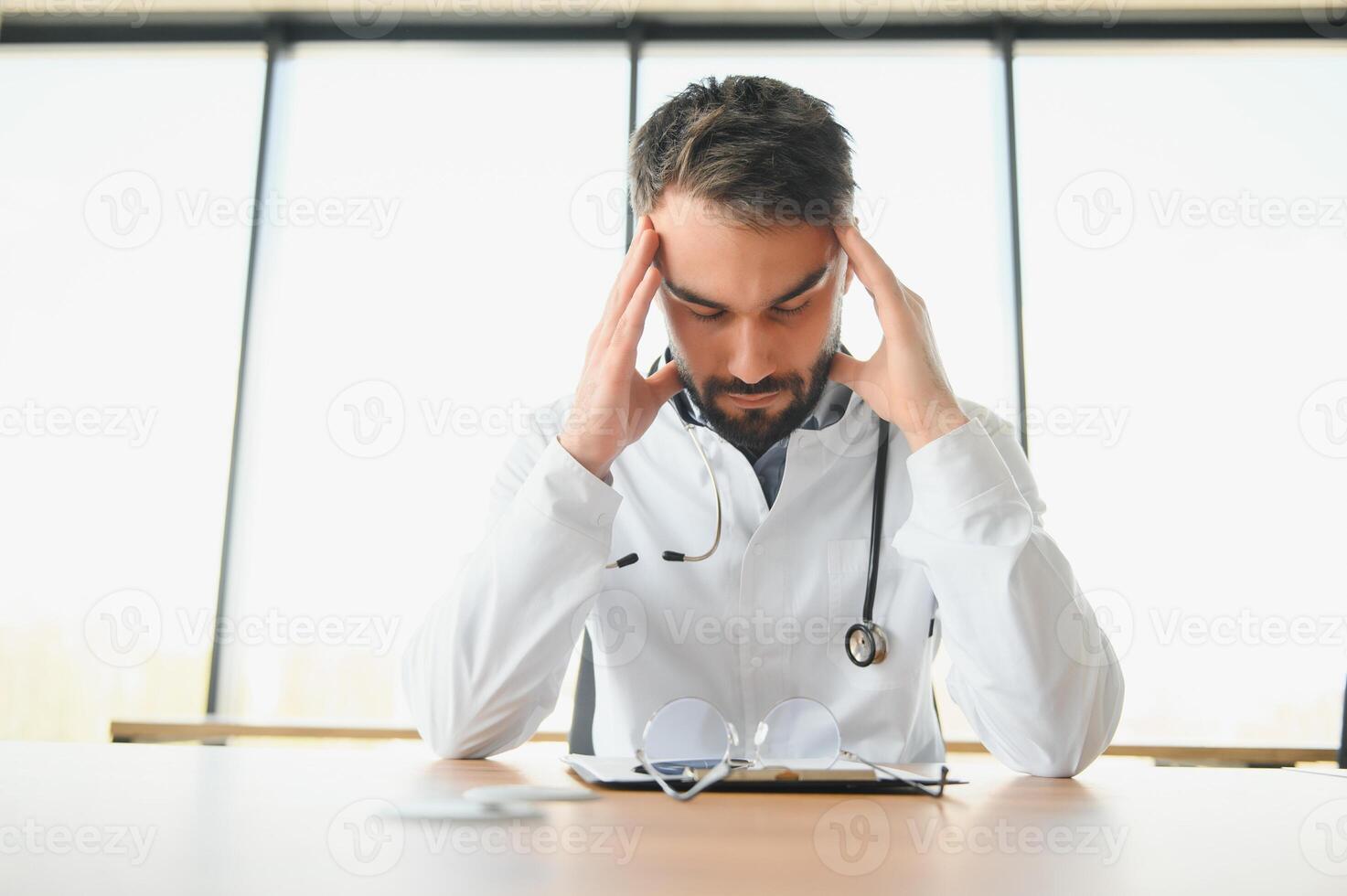 Handsome doctor man wearing medical uniform sitting on his workplace tired holding his head feeling fatigue and headache. Stress and frustration concept photo